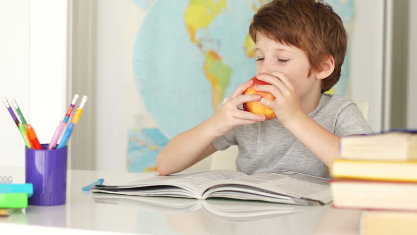 Funny little boy sitting at desk and eating apple