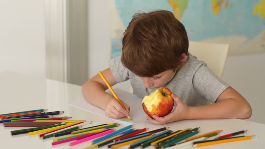 Funny little boy sitting at desk eating apple and drawing