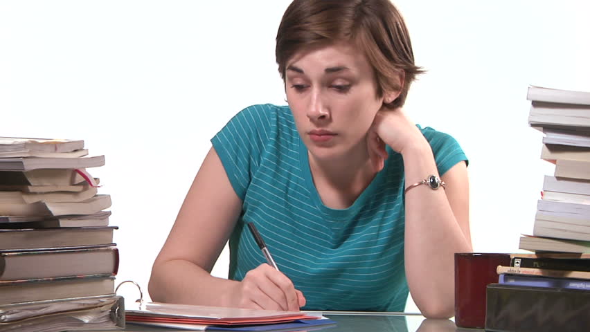 frustrated young woman writes and does homework at a desk piled with books