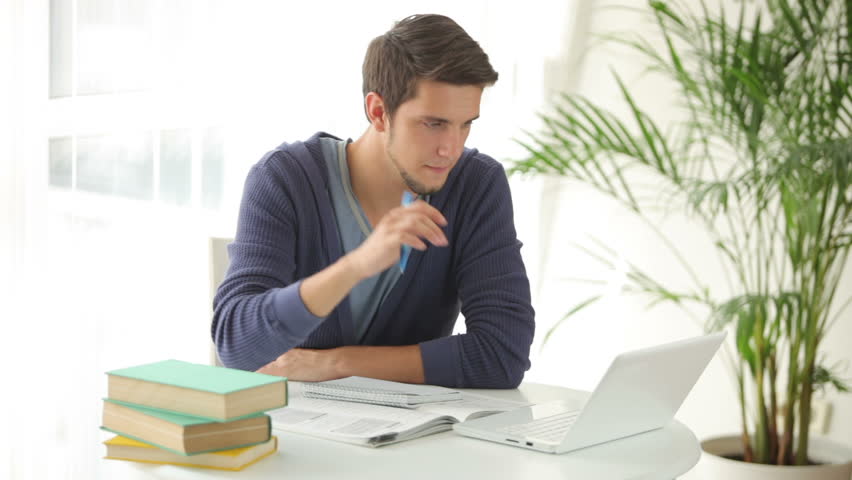 Charming young man studying at table with laptop and books