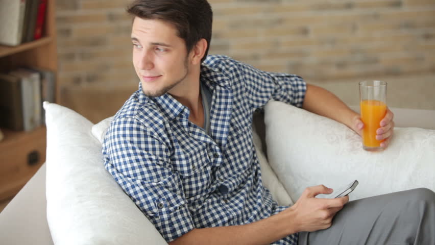 Charming young man sitting on sofa with cellphone and glass of juice