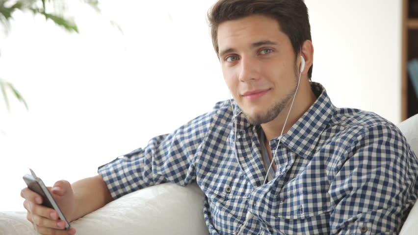 Young man sitting on sofa listening to music with earphones and smiling