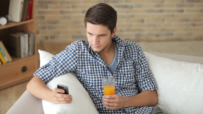 Young man sitting on sofa using cellphone and drinking juice