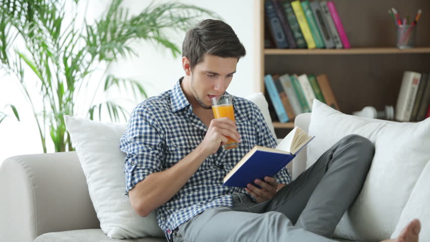 Young man relaxing on sofa reading book and drinking juice