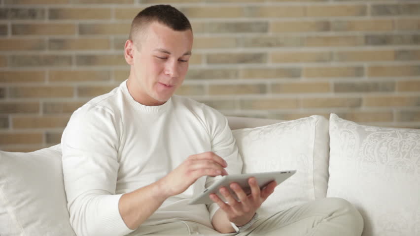 Cheerful guy relaxing on sofa using touchpad and smiling at camera