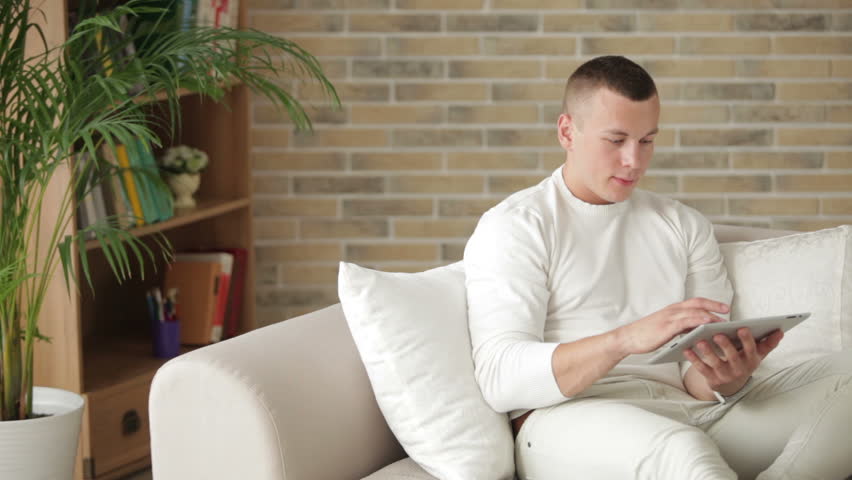 Atttractive guy sitting on couch with touchpad and smiling at camera