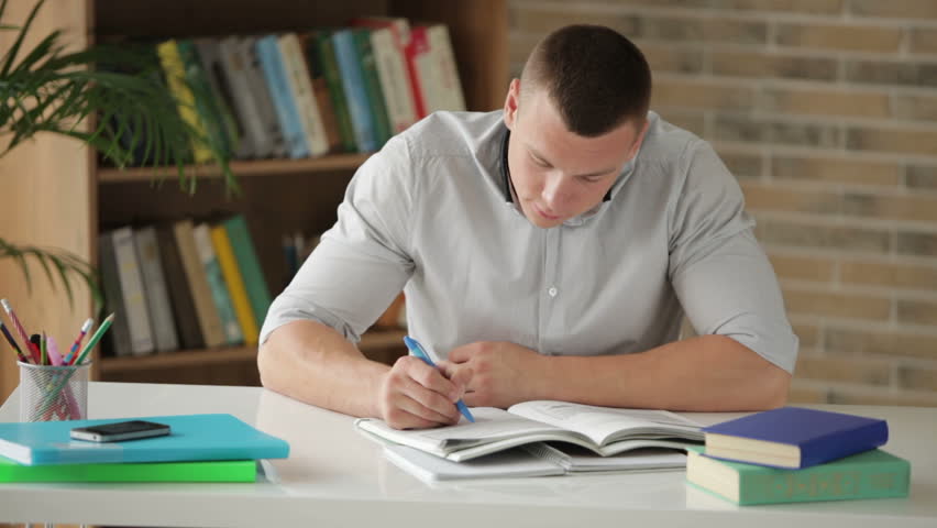Cheerful male student sitting at table and studying with books