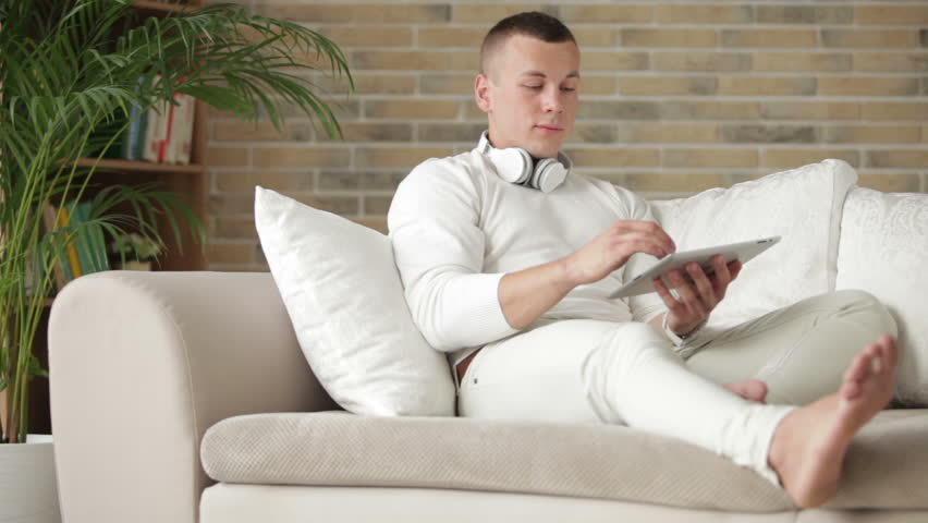 Attractive guy relaxing on sofa with touchpad and smiling at camera