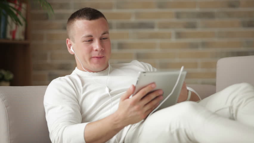 Cheerful guy in earphones relaxing on sofa with touchpad and smiling