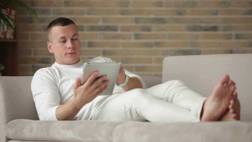 Cheerful guy lying on sofa using touchpad and looking at camera with smile