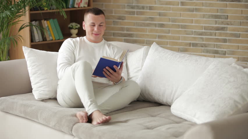 Attractive young man sitting on sofa with book closing it and smiling