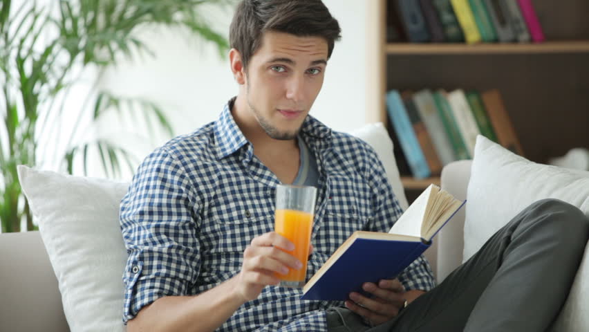 Handsome guy sitting on sofa reading book and drinking juice