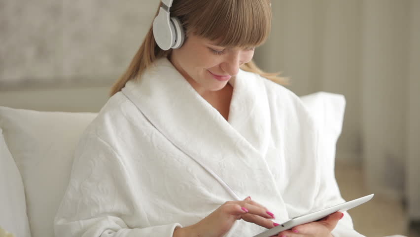 Young woman in headphones sitting on bed and using touchpad