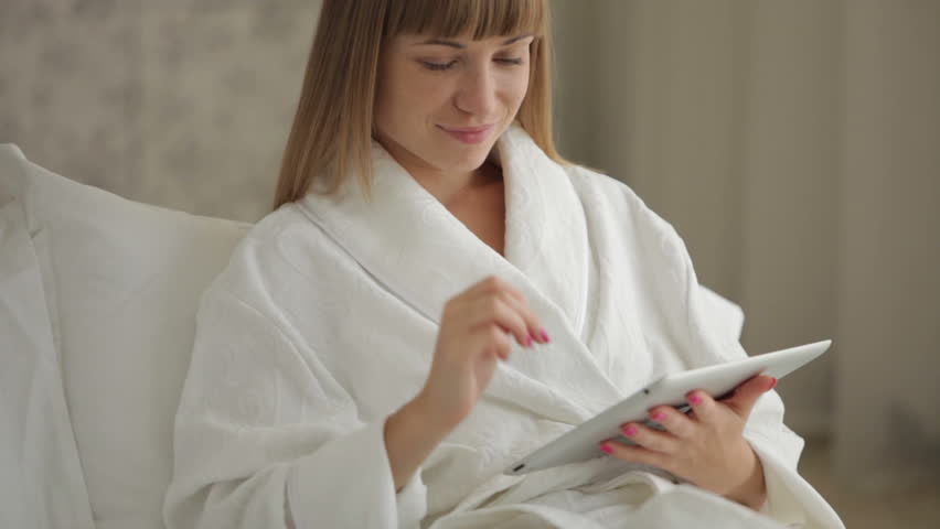 Young woman in bathrobe sitting on bed and using touchpad