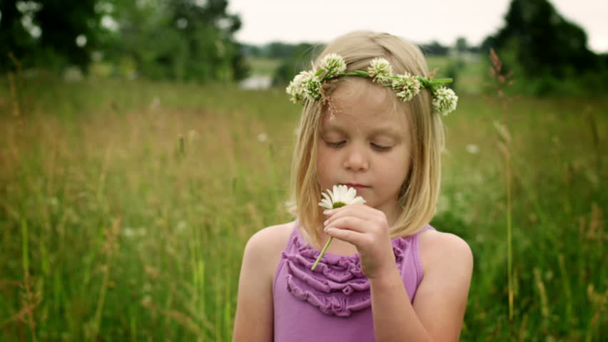 A cute young girl with a flower crown looks to the camera and smells a flower