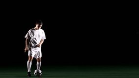 A soccer, or football, player that is dramatically and artistically lit, on an artificial field pitch on a black background, kicks up and juggles a ball with his feet in slow motion wide shot