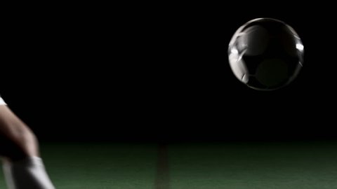 A soccer, or football, player that is dramatically and artistically lit, on an artificial field pitch on a black background, kicks a ball away from the camera at a medium height