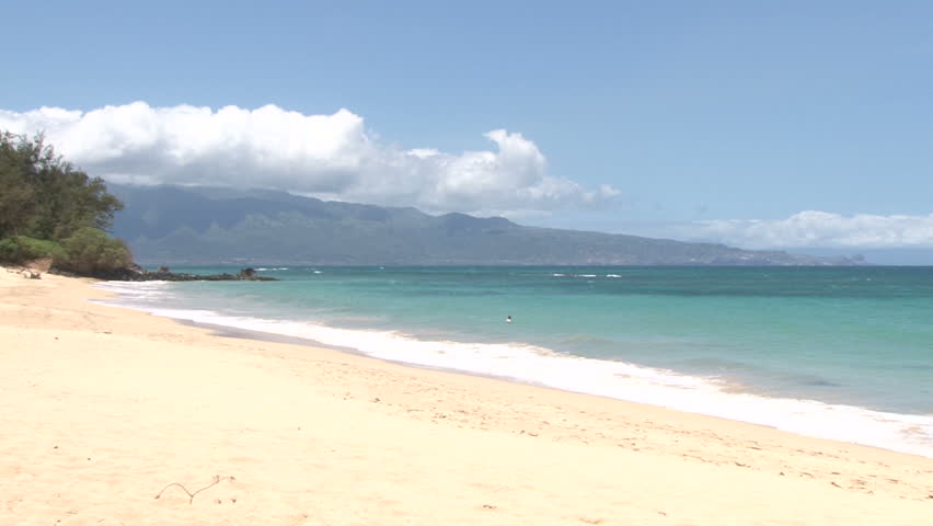 Long camera zoom out on waves crashing gently on quiet sandy beach in Maui,