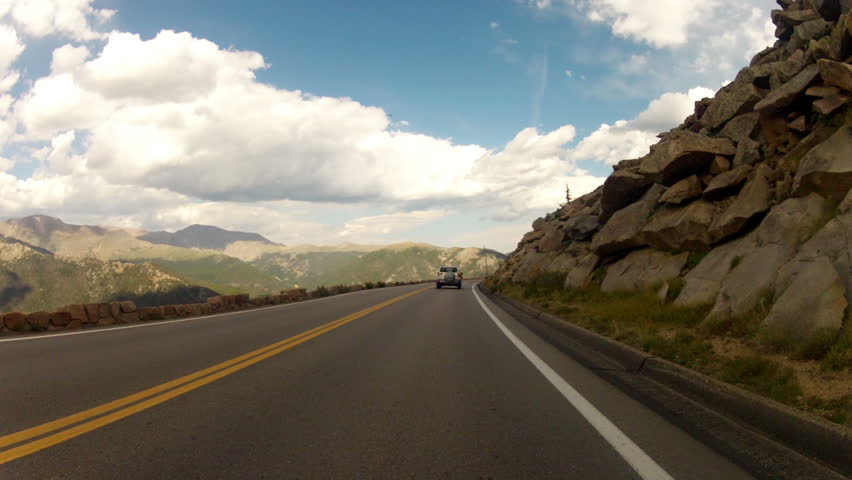 Cliff on one side and boulder strewn mountainside on the other as a Rocky Mountain National Park road unfolds in front of the camera. Car mounted camera footage.