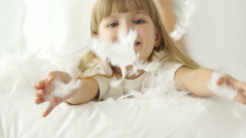 Cute little girl lying in bed laughing and playing with pillow feathers