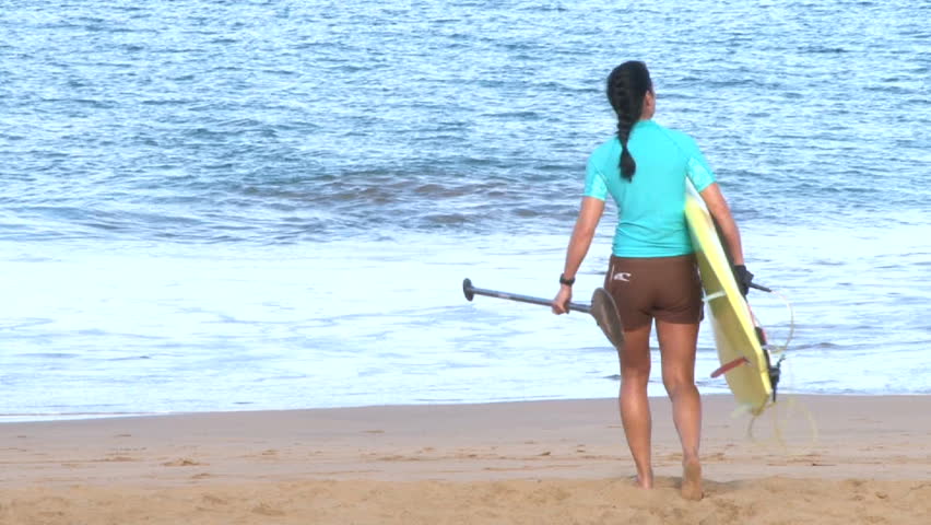 Woman walking towards beach carrying paddle board, getting ready.