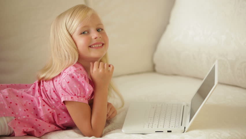 Cheerful little girl lying on sofa using laptop and smiling