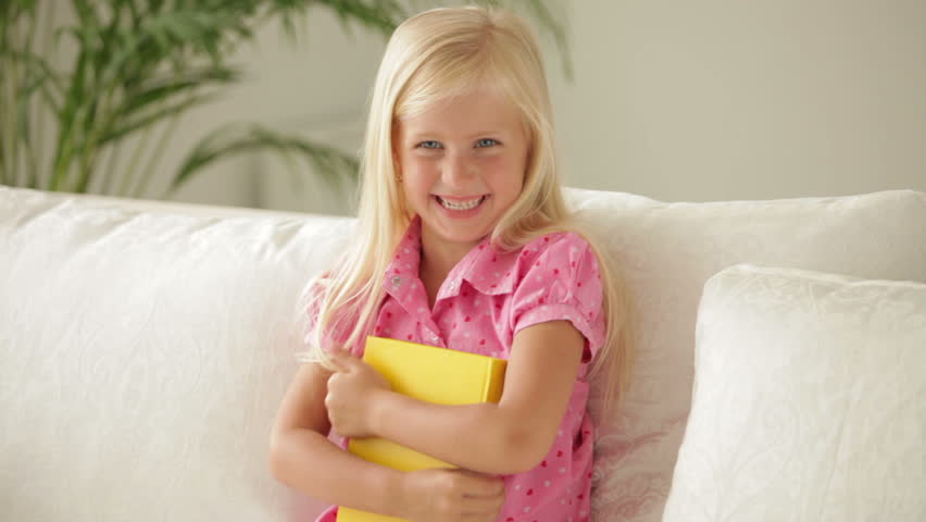 Beautiful little girl sitting on sofa holding book and smiling at camera