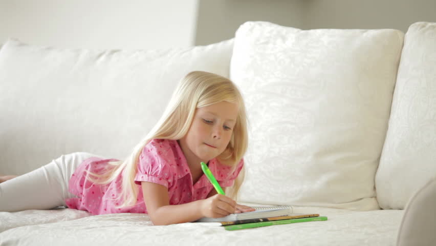 Cute little girl lying on couch writing in notebook and smiling