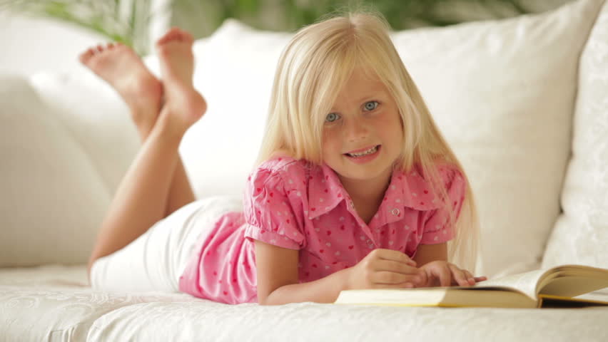 Beautiful little girl lying on sofa reading book and smiling at camera