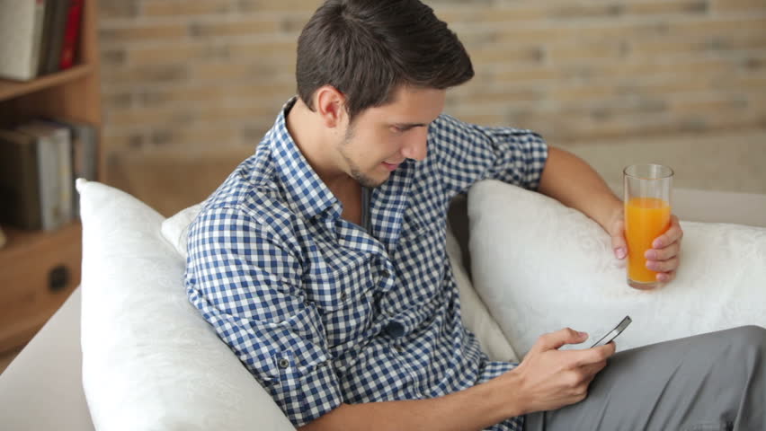 Attractive guy sitting on sofa using cellphone and drinking juice