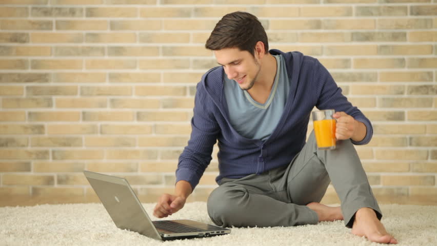 Attractive guy sitting on floor using laptop and drinking juice
