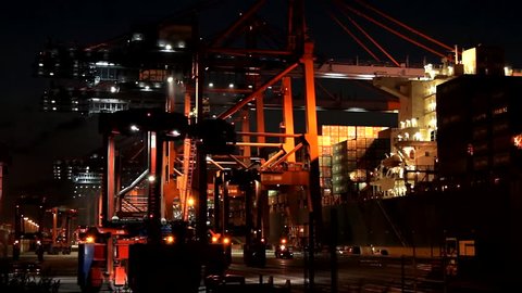 Container ship loading and unloading at night in the port of Hamburg. Waiting vehicles in front with flashing yellow warning lights.
