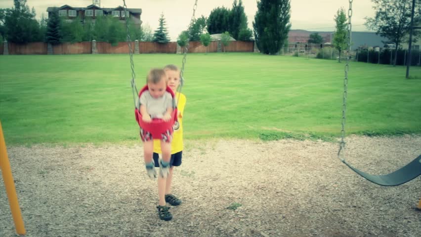 A brother pushing his baby brother on the swings at the park