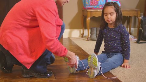 This video has two clips in it, in one a cute little girl tries to tie her shoe alone, in the other her mom pops in and helps to tie it. Two clips put together. Slow motion medium shot.
