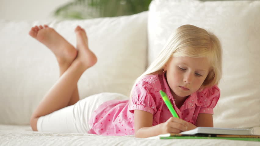 Cute little girl lying on sofa writing in notebook and smiling