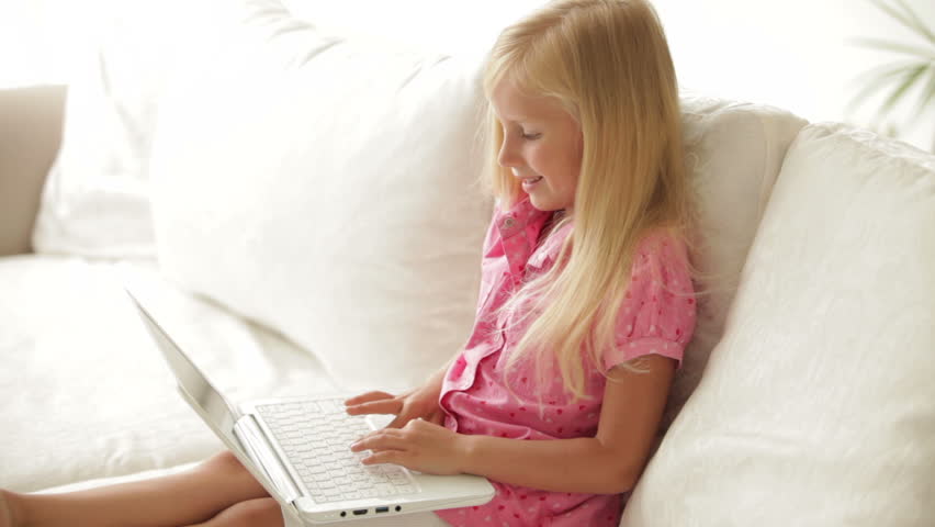 Cute little girl sitting on sofa with laptop and smiling at camera