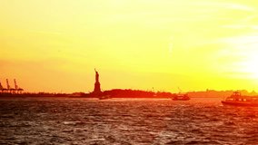 The sun sets over the Statue of Liberty, with boats going by in the water. Wide shot.