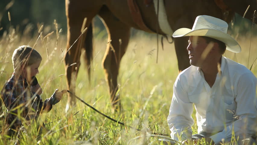 Father and son cowboys sit in grass taking break