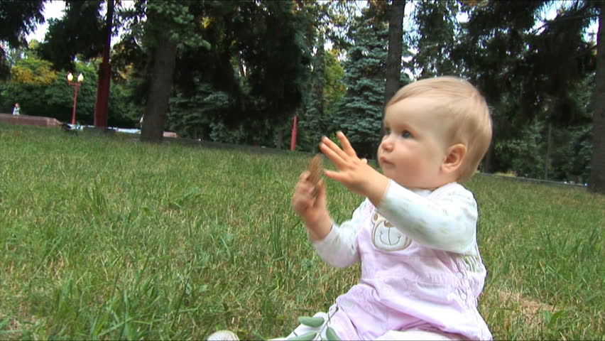 Baby sits on a grass in park. In hands holds a fir cone. Camera-flies