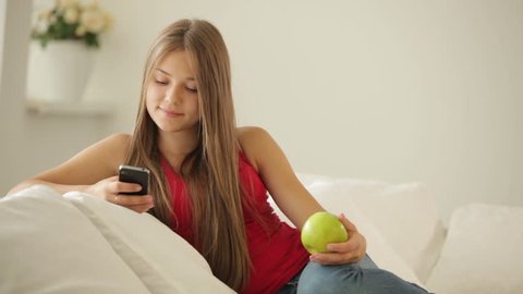 Pretty girl sitting on sofa holding apple and using cellphone