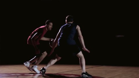 Two basketball players go one on one trying with the red jersey trying to score on the blue jersey in the shadows