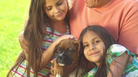 A father and his little girls sit in a park with a boxer puppy. Medium shot.