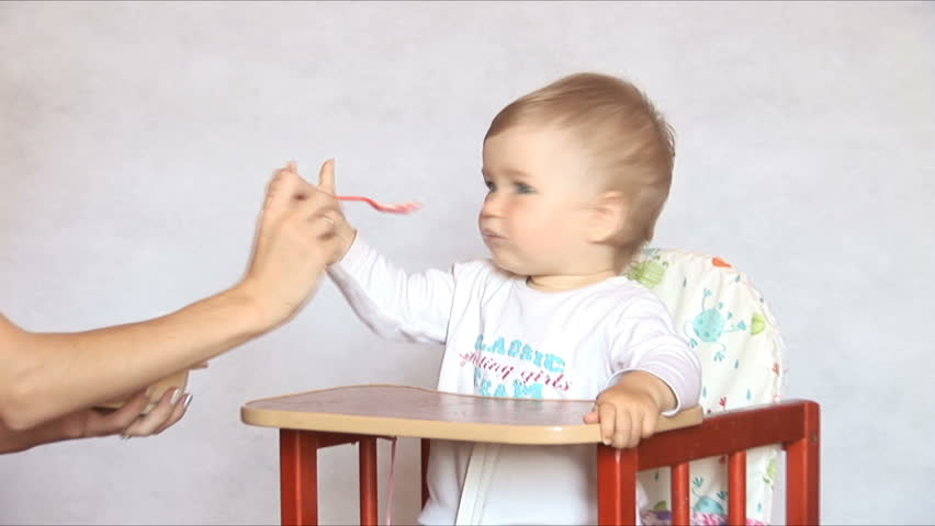 Baby sits at a children's little table. Mum feeds the child with porridge.