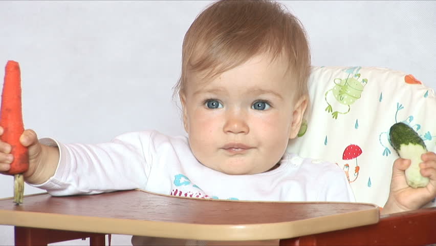 Baby sits at a table and eats a cucumber and carrot. Isolated on the white