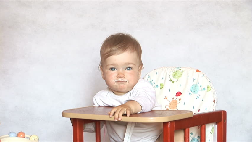 Baby sits at a children's little table. Mum feeds the child with porridge.