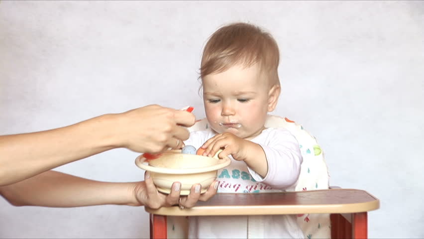 Baby sits at a children's little table. Mum feeds the child with porridge.