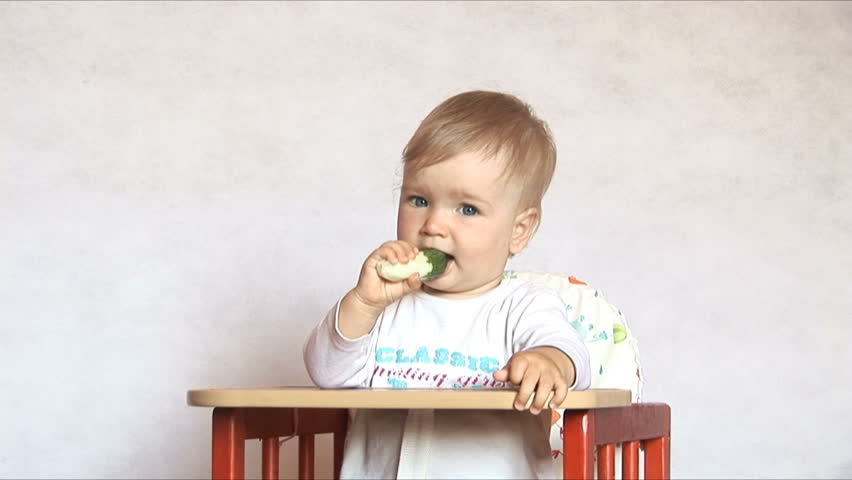 Baby sits at a table and eats a cucumber. Isolated on the white