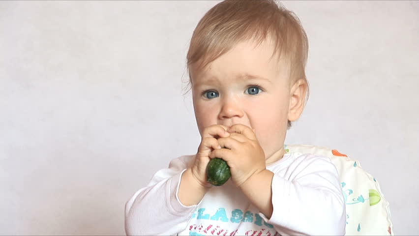 Baby sits at a table and eats a cucumber. Isolated on the white