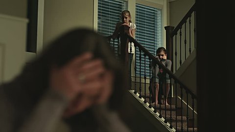 A sad mother sits at the table while her daughters look at her from the stairs. Medium to close up shot.