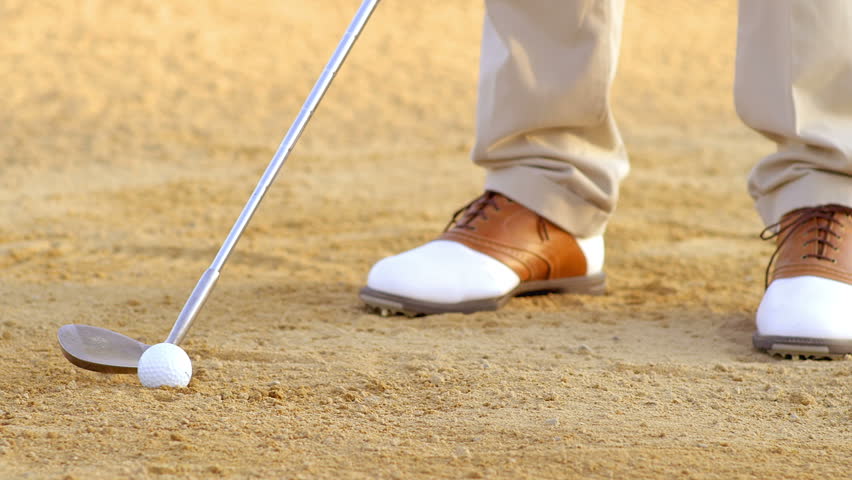 A golfer takes a swing at a golf ball while in a sand trap. Close up slow motion shot.